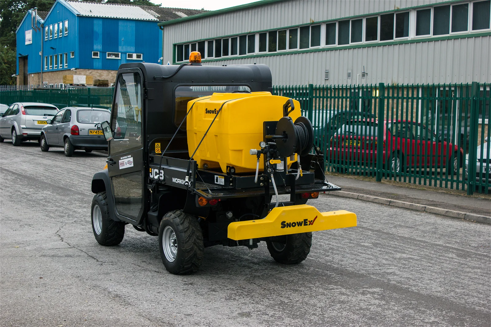 Wessex Snowex Liquid De-Icing System | Vss-1000 | Middlewich Machinery Wessex snowex liquid de-icing system | vss-1000 is a small utility vehicle featuring a yellow spreader attachment, is parked on a paved road near a fenced industrial building. Several parked cars are visible in the background.