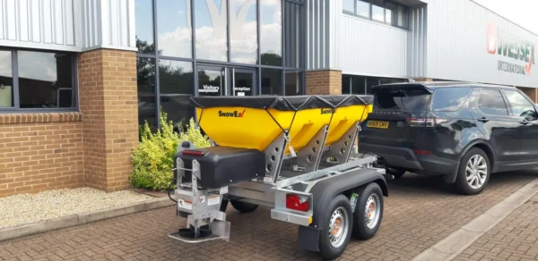 A yellow wessex snowex helixx spreader on a dual-axle trailer is hitched to a black suv parked outside a commercial building.